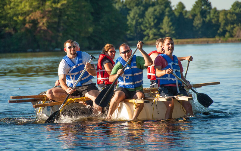 A group of six people are on a makeshift raft in the water. They are using paddles to propel themselves forward. The raft appears to be constructed from barrels and wooden planks. The people are wearing life jackets, and the atmosphere seems cheerful and adventurous. The water is calm, and the sun is shining, suggesting a pleasant day for a water activity.